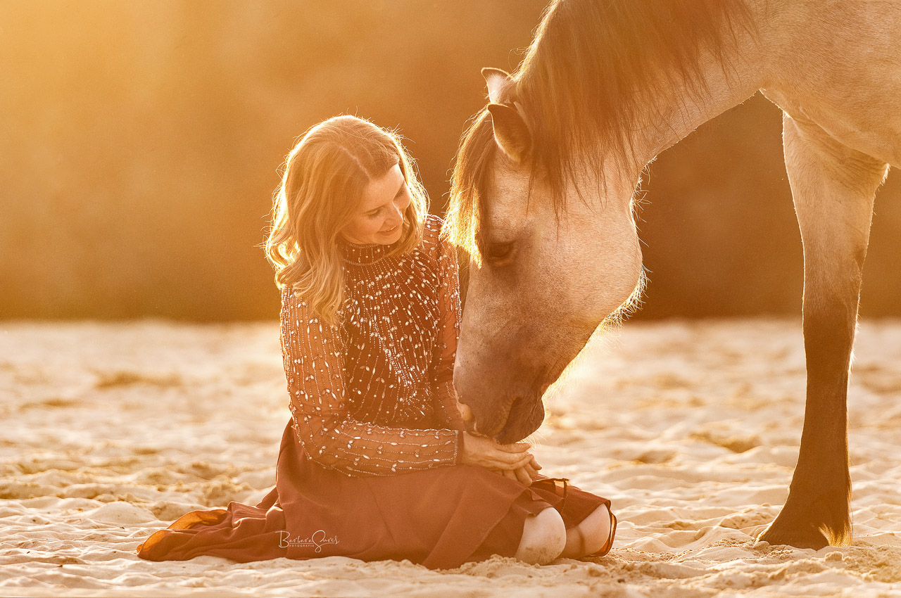 Eine junge Frau sitzt im Sand und ihr Pferd neigt sich zu ihr