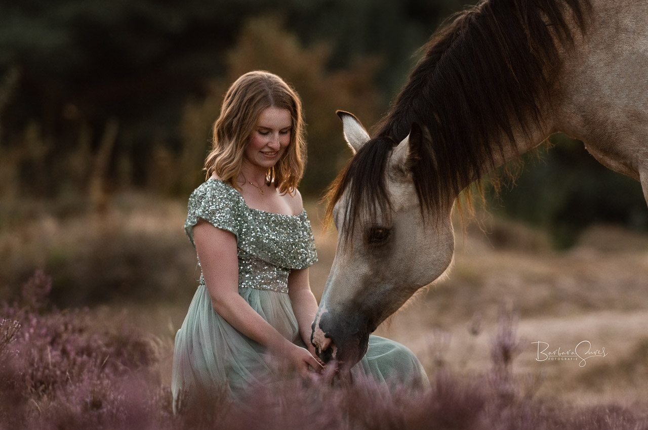 Eine junge Frau hockt in der Heide vor ihrem Pferd